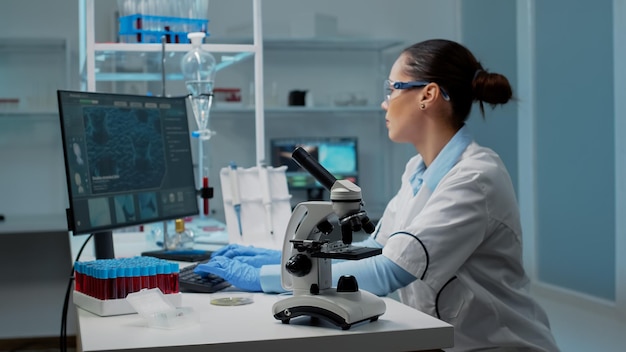 Onderzoekende arts in laboratorium met behulp van chemische hulpmiddelen voor onderzoek. Wetenschappervrouw typt op de computer en kijkt naar bacteriën in een petrischaal terwijl ze professionele apparatuur op het bureau heeft