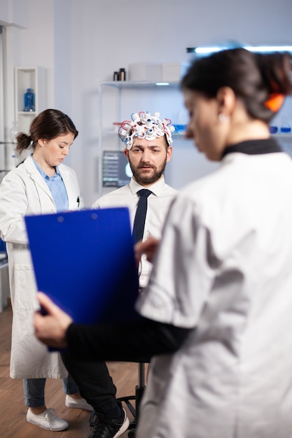 Onderzoeker neuroloog arts die de ziektesymptomen van de mens vraagt en naar het klembord kijkt voordat de hersenscan wordt uitgevoerd met een headset voor het scannen van hersengolven. Wetenschapper die gezondheidsstatus, zenuwstelsel, tomografiescan analyseert.
