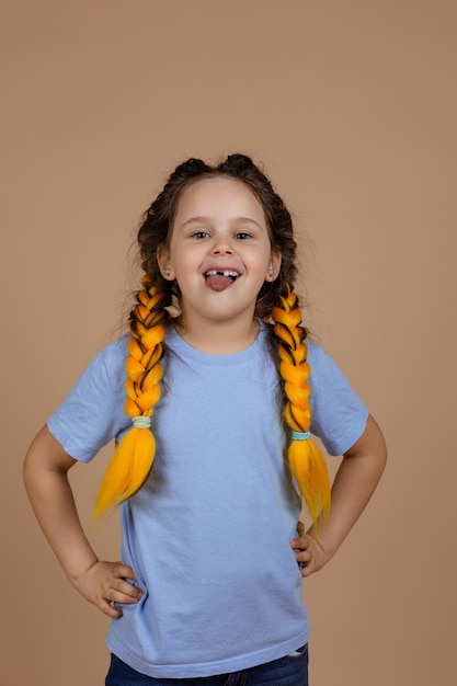 Ondeugend klein glanzend meisje met een lichte glimlach die tong toont die naar camera kijkt die zich misdraagt met kanekalon-vlechten op beige achtergrond met blauwe tshirt slechte gewoonten