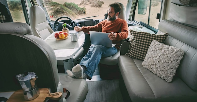 One alone man sitting and relaxing inside a camper van alternative home vanlife lifestyle off grid using a laptop and taking a coffee in total relax freedom Indoor motor home travel leisure activity