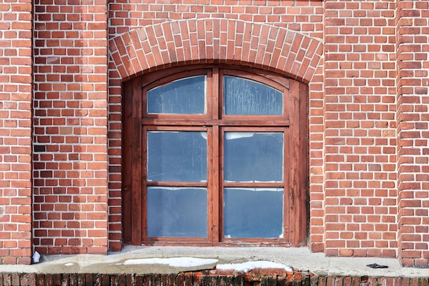 One arched glass window on old red brick wall. Vintage window in brown wooden frame on red brick wall of industrial building.