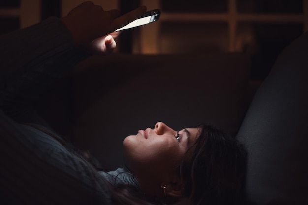 Одна красивая молодая женщина держит и пользуется телефоном дома поздно ночью на диване Девушка-подросток болтает с друзьями в сети онлайн Наслаждаясь технологиями и интернетом