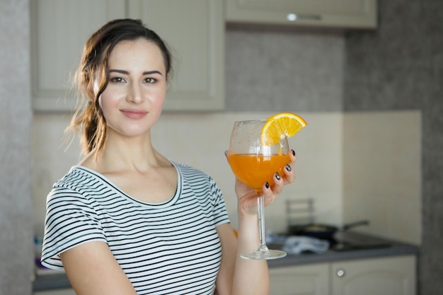 Одна красивая молодая женщина в полосатой футболке держит в руке стакан с долькой апельсина и соком на кухне