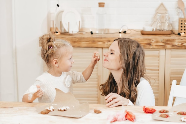 One blonde girl paints her mom's face with cream. Mom and daughter decorate Christmas cookies with icing on a wooden table in the kitchen.