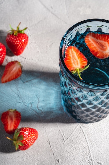 Una tazza di vetro geometrica blu con acqua fresca e frutta fragola con raggi di luce ombreggiati colorati sulla pietra