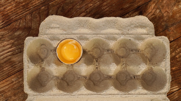 One broken egg in a cardboard egg tray. half egg with yolk in\
an empty box on wooden background