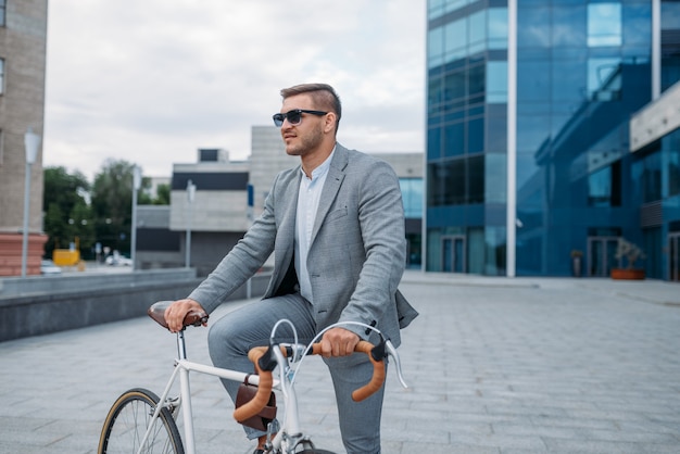 サングラスをかけたあるビジネスマンが、ダウンタウンのオフィスビルで自転車に乗ってポーズをとっています。街の通りでエコ輸送に乗るビジネスパーソン