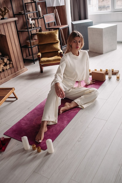 One Caucasian blonde woman in white relaxing on bright pink mat barefoot surrounded by candles ready for meditation at home.