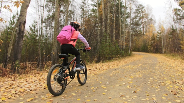 One caucasian children rides bike road in autumn park little\
girl riding black orange cycle in forest kid goes do bicycle sports\
biker motion ride with backpack and helmet mountain bike hard\
tail