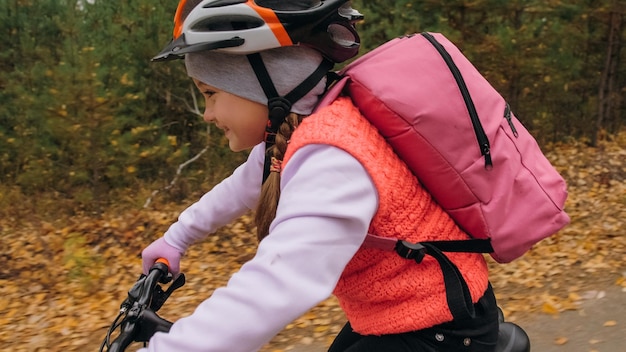 一人の白人の子供たちが秋の公園で自転車の道に乗る森の中でブラックオレンジのサイクルに乗っている小さな女の子子供は自転車スポーツをしますバックパックとヘルメットでバイカーモーションライドマウンテンバイクハードテール