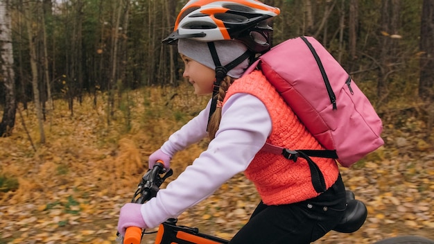 一人の白人の子供たちが秋の公園で自転車の道に乗る森の中でブラックオレンジのサイクルに乗っている小さな女の子子供は自転車スポーツをしますバックパックとヘルメットでバイカーモーションライドマウンテンバイクハードテール