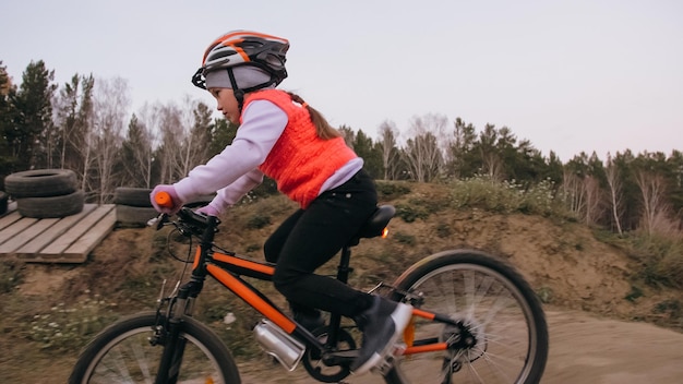 One caucasian children rides bike road track in dirt park Girl riding black orange cycle in racetrack Kid goes do bicycle sports Biker motion ride with backpack and helmet Mountain bike hard tail