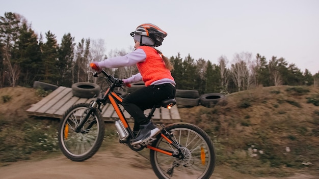 One caucasian children rides bike road track in dirt park girl\
riding black orange cycle in racetrack kid goes do bicycle sports\
biker motion ride with backpack and helmet mountain bike hard\
tail