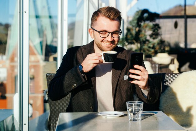 One caucasian man wearing glasses holds smartphone and drinks coffee from cup at cafe. Remote work via smart phone. City life. Business person at break. Casual fashion. Success concept. Mixed media.