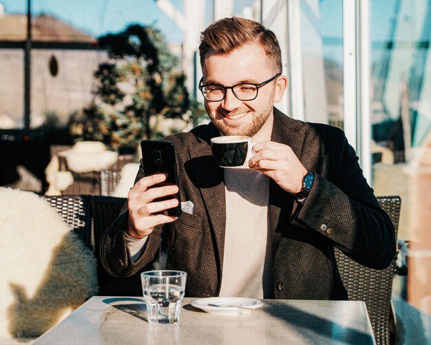 眼鏡をかけている一人の白人男性がスマートフォンを持って、カフェでカップからコーヒーを飲みます。スマートフォンを介したリモートワーク。街の暮らし。休憩中のビジネスパーソン。カジュアルファッション。成功のコンセプト。ミクストメディア。