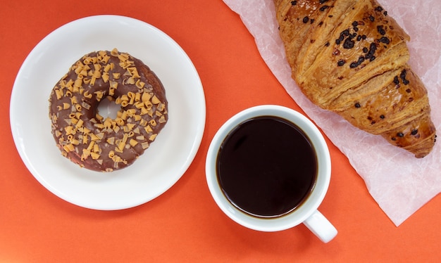 Один шоколадный пончик, круассан и черный кофе американо без молока в белой чашке на ярком фоне. Вид сверху, плоская планировка. Свежезаваренный или растворимый горячий кофейный напиток со сладкой выпечкой.