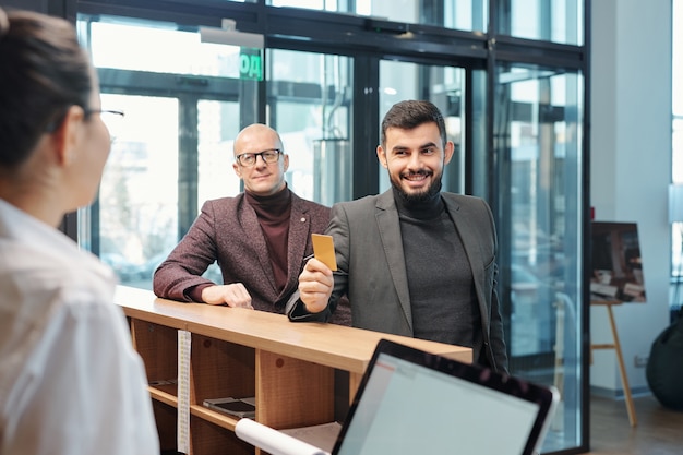 One of contemporary business travelers showing plastic card to hotel receptionist while ordering room for night