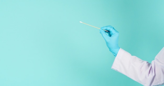 One cotton stick for swab in hand with doctor gown and blue\
medical gloves or latex glove on mint green or tiffany blue\
background.covid-19 concept