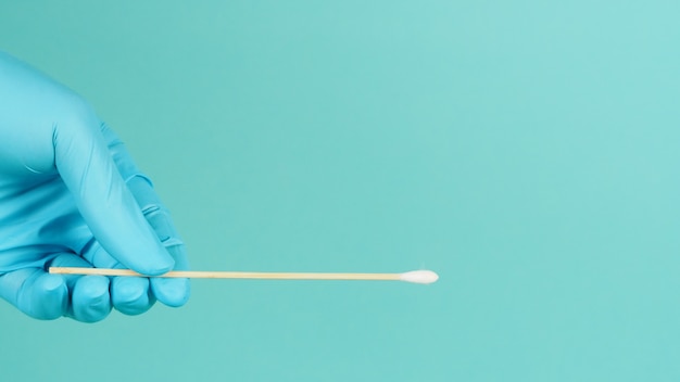 One cotton stick for swab test in hand with blue medical gloves\
or latex glove on mint green or tiffany blue background.covid-19\
concept. close up