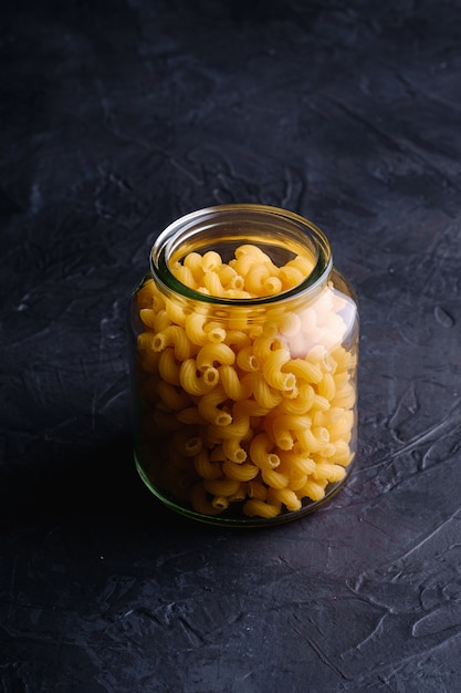 One glass jar with cavatappi uncooked golden wheat curly pasta on textured dark black surface, angle view