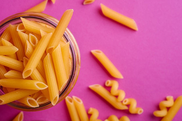 Un barattolo di vetro con la pasta dorata cruda del tubo del grano del penne sulla tavola rosa minima, macro di vista superiore