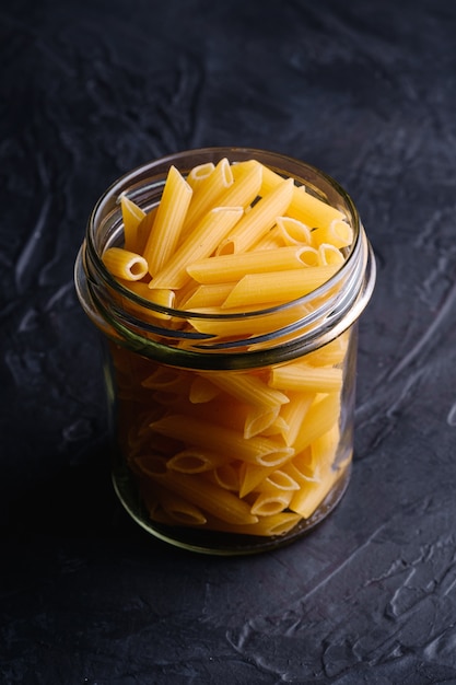 One glass jar with penne uncooked golden wheat tube pasta on textured dark black table, angle view