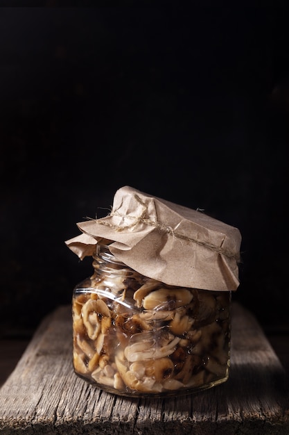 One glass jar with pickled forest mushrooms honey agarics under paper cover on old wooden board.