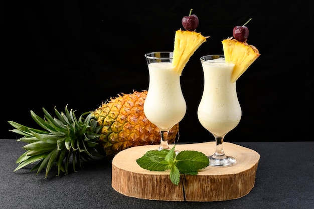 One glasses with pina colada cocktail a whole pineapple next to the drinks on black background