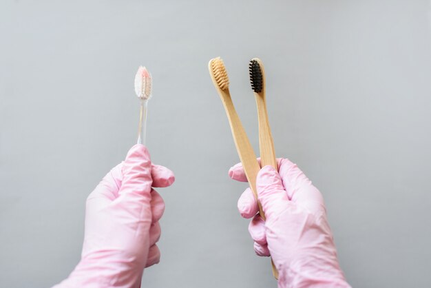 In one hand a plastic brush for teeth, in the other two bamboo brushes for teeth. Hands in pink gloves hold brushes.