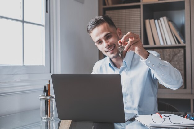 One happy caucasian man smiling and talking with clients online in video conference showing them their house keys. Busy worker using laptop or computer working.