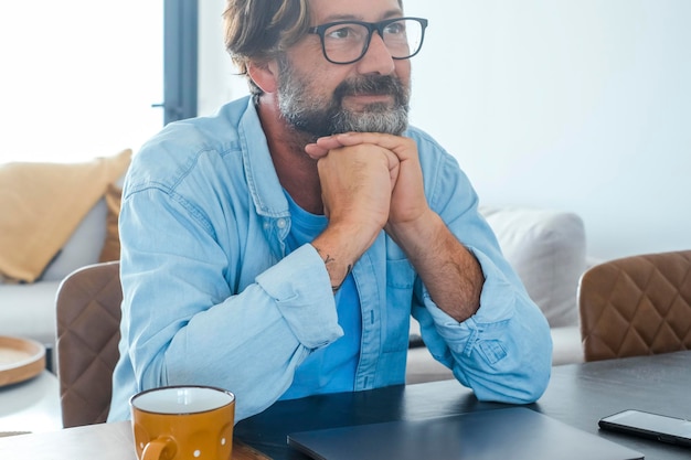 One happy and serene man looking up the air at home with closed laptop on the table. Concept of dreamer and daydreaming people. Relax after online work. Adult relaxing in front of a computer shut down