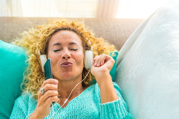 One happy and young woman singing and listening music on the couch or sofa at home with white headphones - music theraphy lifestyle