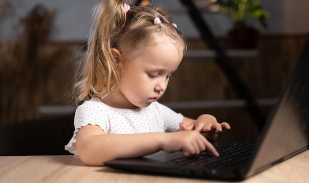 One little preschool girl in the evening sits at home at a table with a laptop with her fingers focused and is typing text