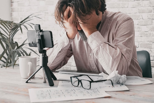 One man desperate holding head with hands in front of a desk full of business documents and phone on a tripod for video call. Online business manager problems and crisis. Bankrupt concept moment.