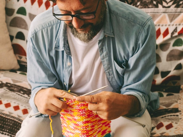 One man at home busy in new hobby activity doing knitting work with wool and needles Single male mature in indoor leisure activity alone People in alternative work Sitting on sofa in apartment