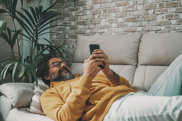 One mature man use mobile phone cellular laying on tne sofa at home in relax leisure online activity People using cell smartphone indoor in apartment Concept of real life and surf the web alone