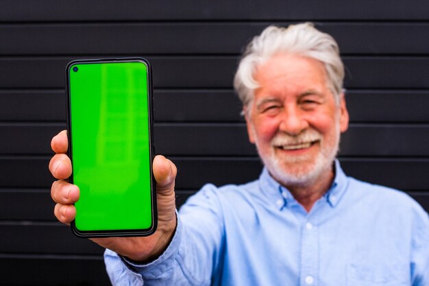 One old and mature senior man holding a mobile phone with green screen and showing it to the camera