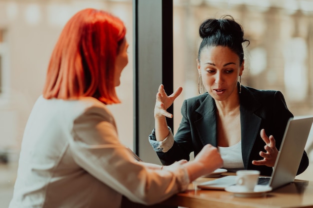 One-on-one meeting.Two young business women sitting at table in cafe. Girl shows colleague information on laptop screen. Girl using smartphone, blogging. Teamwork, business meeting. Freelancers workin