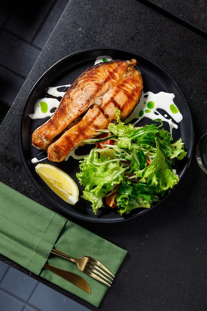 Un pezzo di salmone al forno grigliato pepe limone e sale su un piatto nero con foglie di lattuga su uno sfondo di pietra