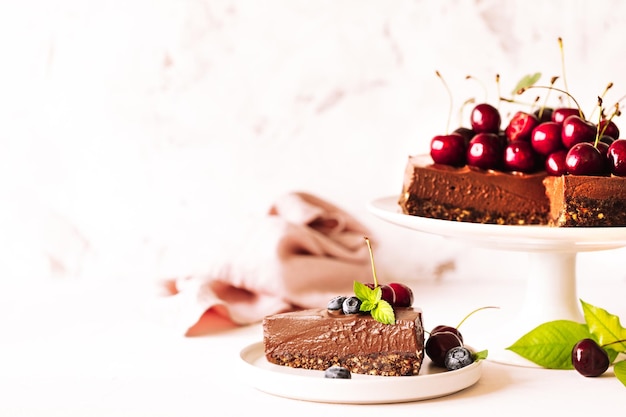 One piece of chocolate cheesecake decorated with fresh cherries blueberries and a whole pie on a ceramic stand on a beige background Horizontal Copy Space