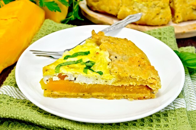 Один кусок пирога из тыквы, соленого сыра фета, яиц, сливок и зелени в тарелке на полотенце, базилик на темной деревянной доске