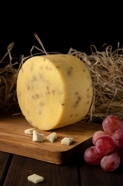 One piece of round yellow hard cheese with sunflower seeds, hay, branches of red and green grapes on a cutting board side view