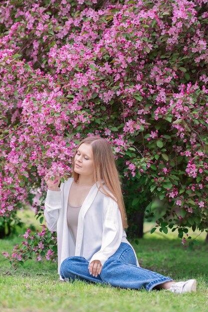 Foto una bella giovane bionda seduta sotto l'albero in fiore con fiori rosa che indossa una camicetta bianca