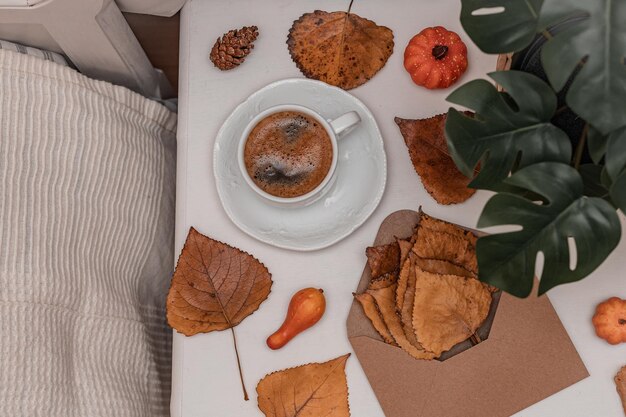 One small mug of coffee with a saucer a envelope pumpkins and a flower on a white bedside table