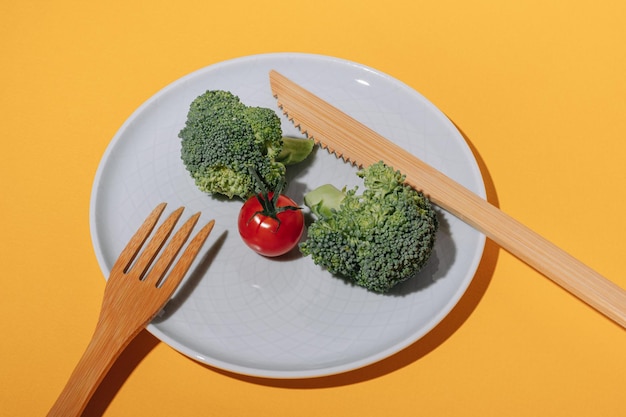 One small red cherry tomato and broccoli on a plate and bamboo fork and knife on a yellow background