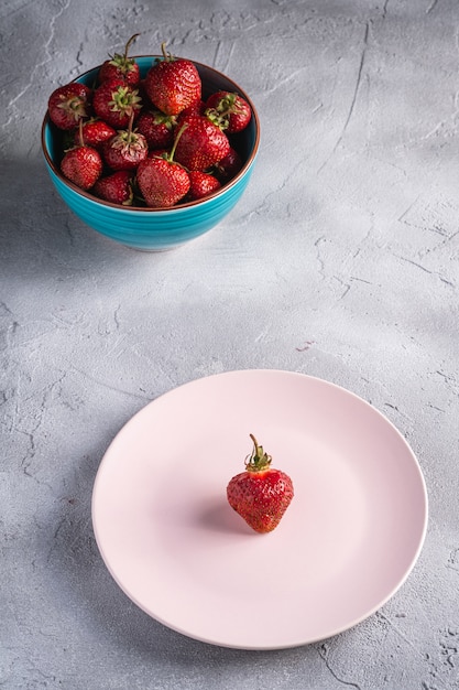 One strawberry in pink plate near to fresh ripe strawberries fruits in blue bowl