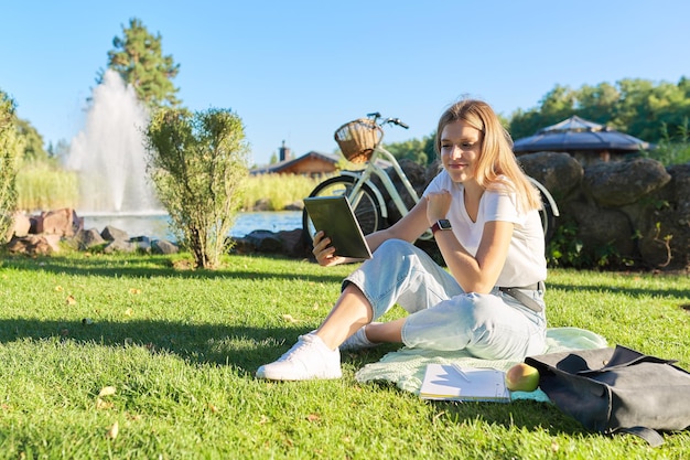 공원 연구에서 녹색 잔디 잔디밭에 노트북 디지털 태블릿을 들고 앉아 있는 한 10대 여학생은 온라인으로 통신하며 원격으로 강의를 듣습니다.