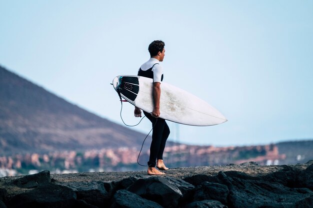 Foto un adolescente che entra in acqua con il suo surftable sotto il braccio pronto per andare a fare surf - atleta attivo va ad allenarsi in acqua - uomo con muta