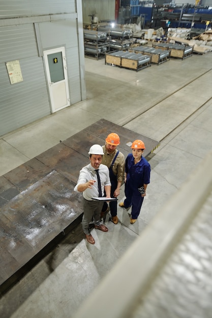 One of three young engineers in uniform and hardhats pointing at new huge industrial machine while showing it to colleagues