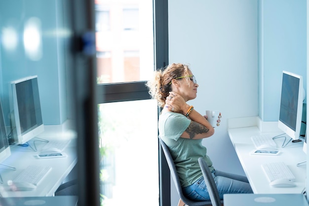 One tired woman worker touch her neck sitting against a desktop computer in office workplace Modern job and painful bad posture Unhealthy job Stressed female people end to work for back ache